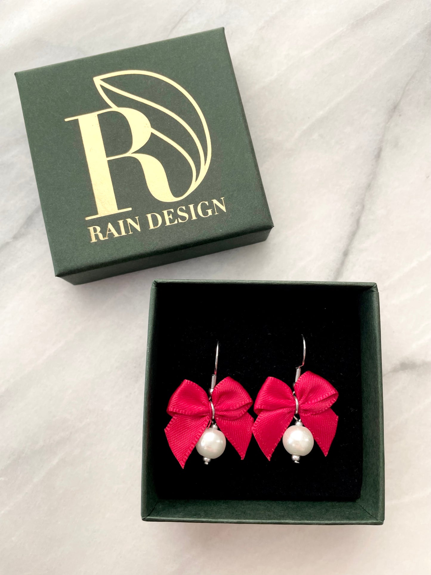 Red bow earrings with white pearls in a 'Rain Design' box on a white fabric background